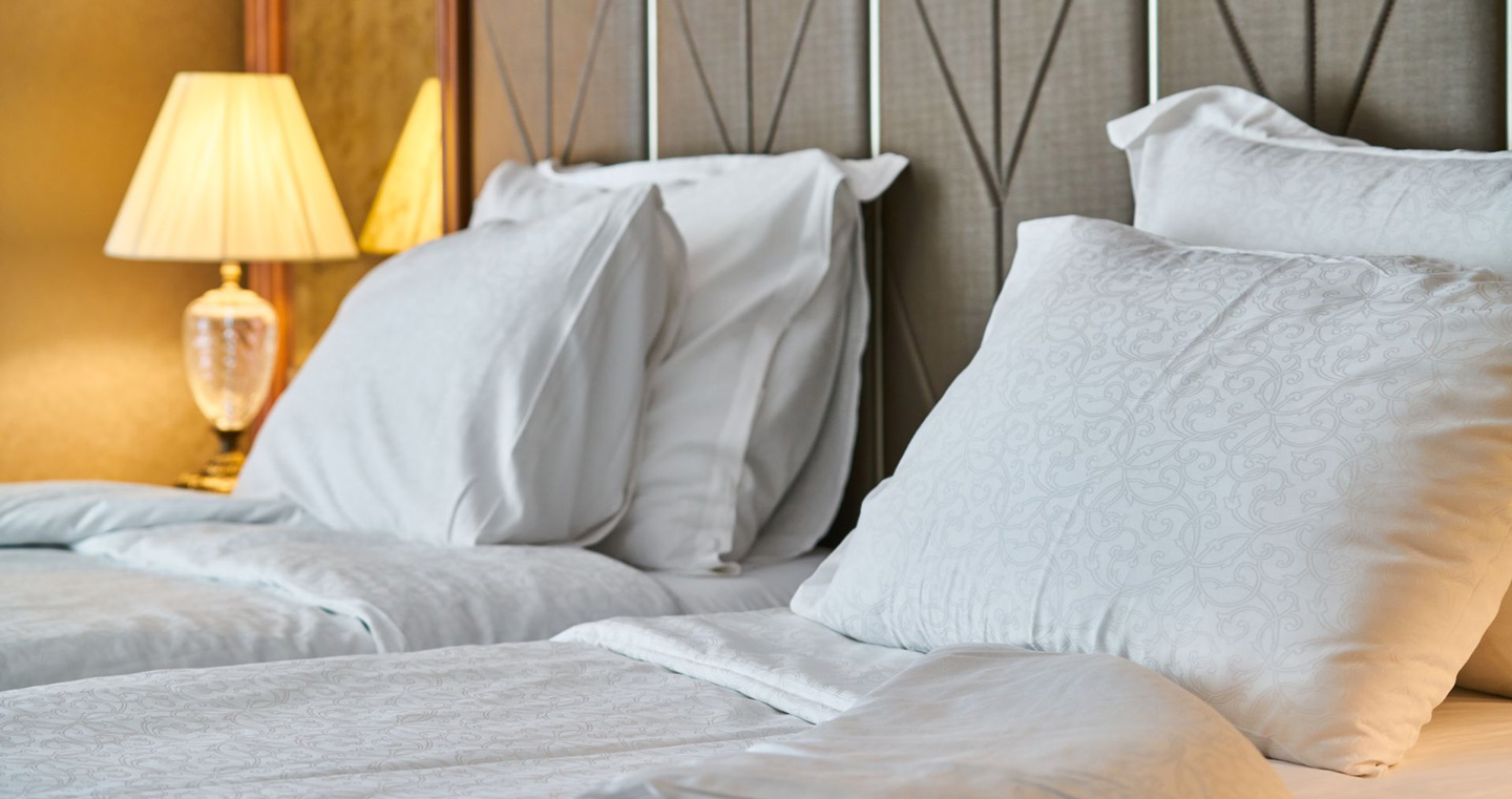 Neatly made bed with white pillows and a headboard beside a lit bedside lamp Neatly made bed with white pillows and a headboard beside a lit bedside lamp