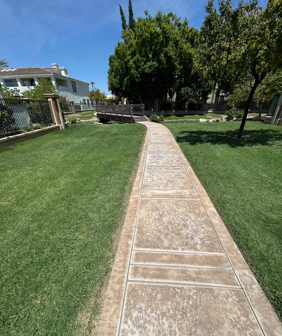 Concrete walkway winding through a mown HOA lawn past shade trees and an iron fence