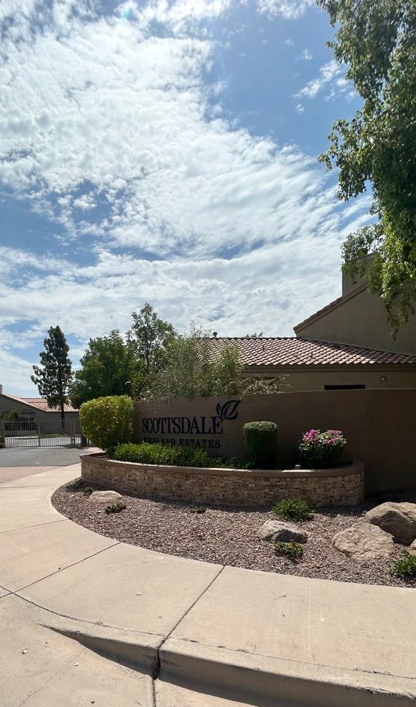 Community entrance sign in a stone-faced planter with flowering shrubs beside a curving sidewalk