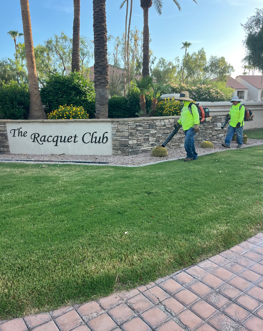 Two TerraPro crew members using backpack blowers at The Racquet Club community entrance sign