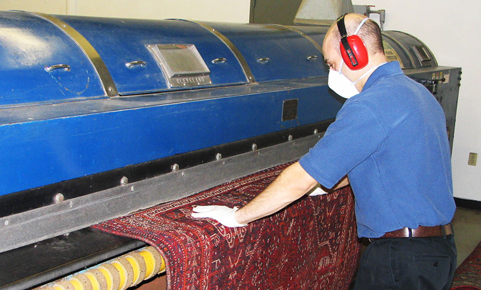 Worker in ear protection feeding a red oriental rug into the blue rug dusting machine