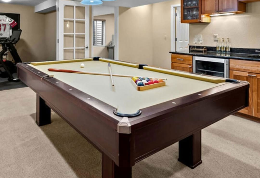 Basement game room with a pool table and cue resting on plush wall-to-wall carpet
