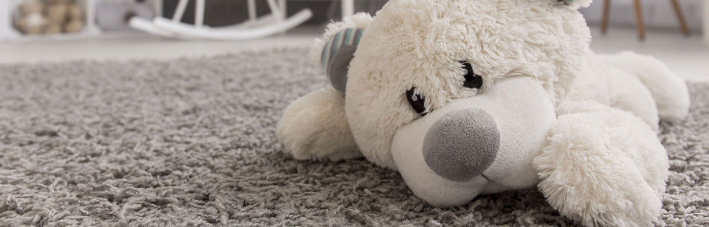 White plush teddy bear lying on soft grey shag carpet in front of a wooden rocking chair