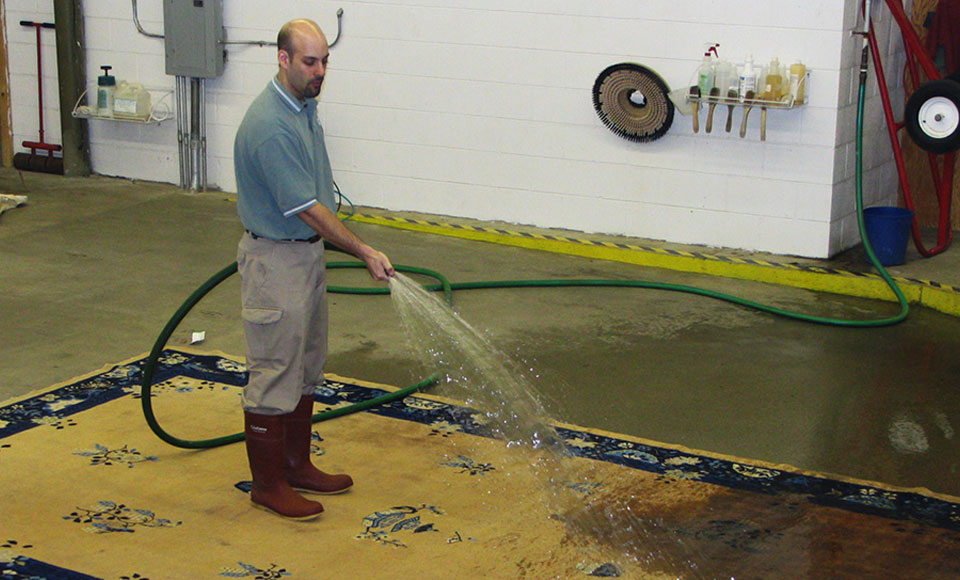 Worker in rubber boots rinsing a soapy area rug with a hose on the plant wash floor